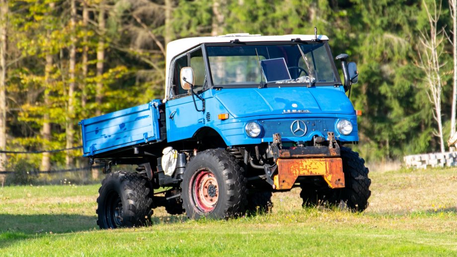 Ein blauer Unimog fährt über eine grüne Wiese, im Hintergrund sind Bäume zu sehen. 