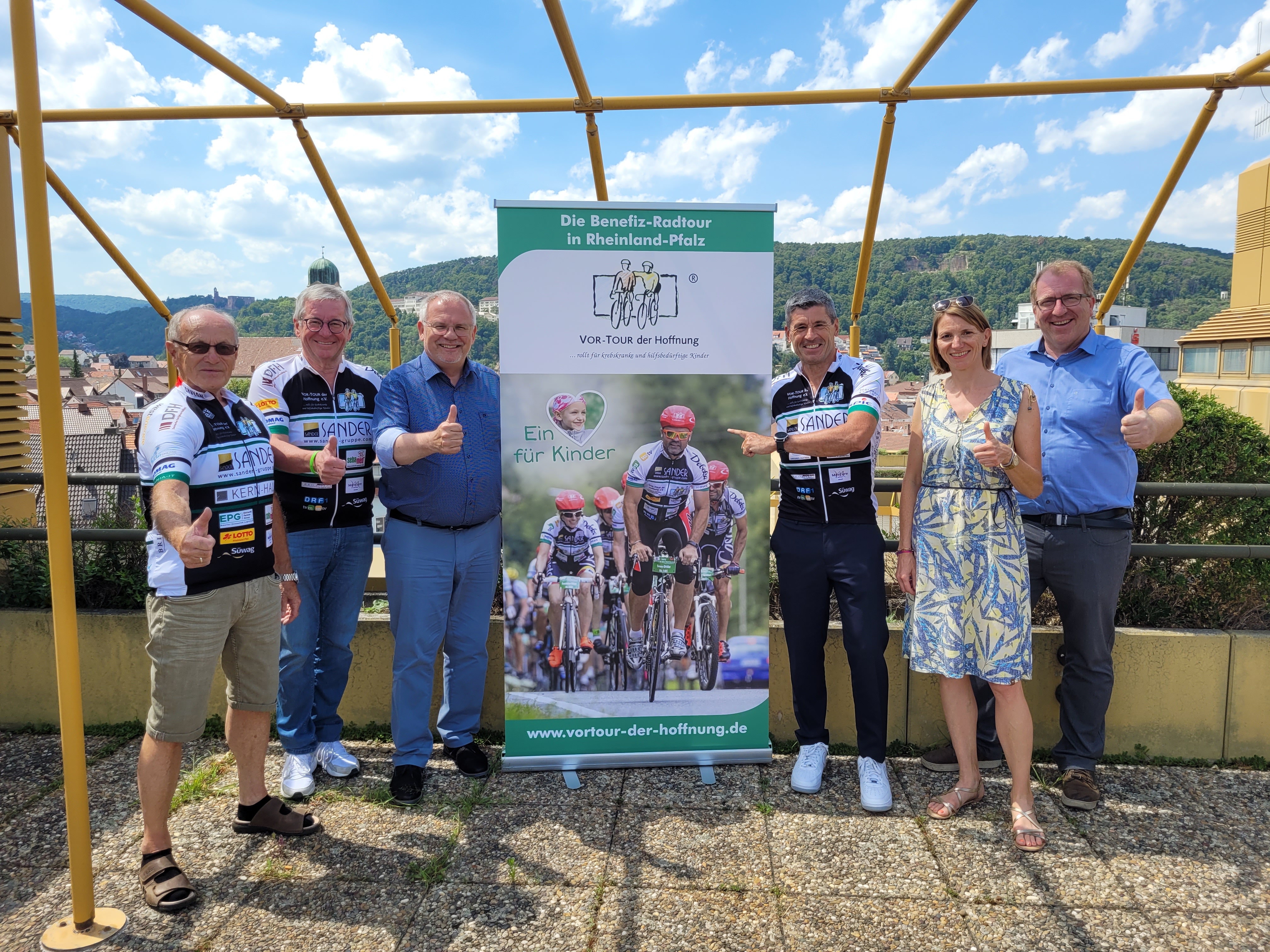 Je drei Männer stehen links und rechts neben einem Werbebanner für die "Vor-Tour der Hoffnung". 