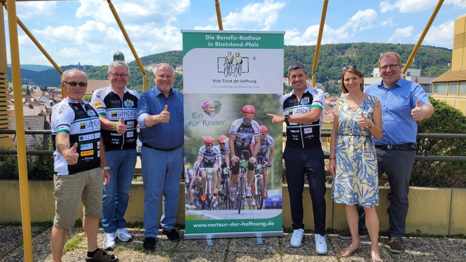 Je drei Männer stehen links und rechts neben einem Werbebanner für die "Vor-Tour der Hoffnung". 