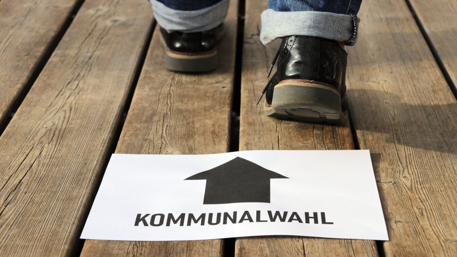 Eine Person mit blauen Jeans und schwarzen Schuhen, von der nur die Füße und Knöchel zu sehen sind, läuft über einen alten Holzdielenboden nach hinten aus dem Bild. Davor liegt ein Papier mit der Aufschrift "Kommunalwahl" und einem Pfeil, der in Laufrichtung zeigt. 