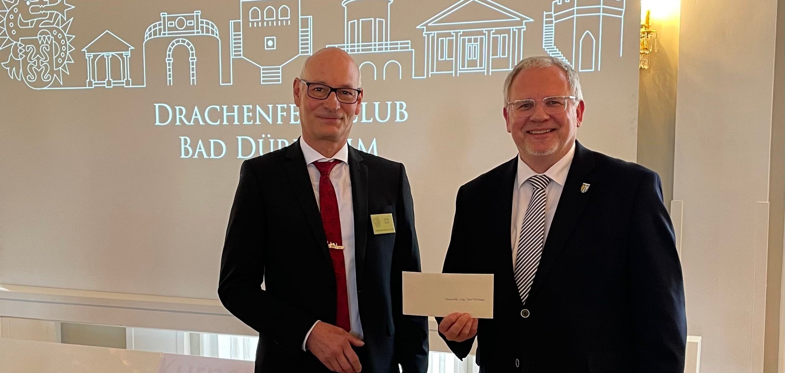 Zwei Männer mit einem weißen Briefumschlag. Im Hintergrund ist auf einer Leinwand der Text "Festakt zum 150-jährigen Bestehen des Drachenfelsclub Bad Dürkheim" zu lesen.