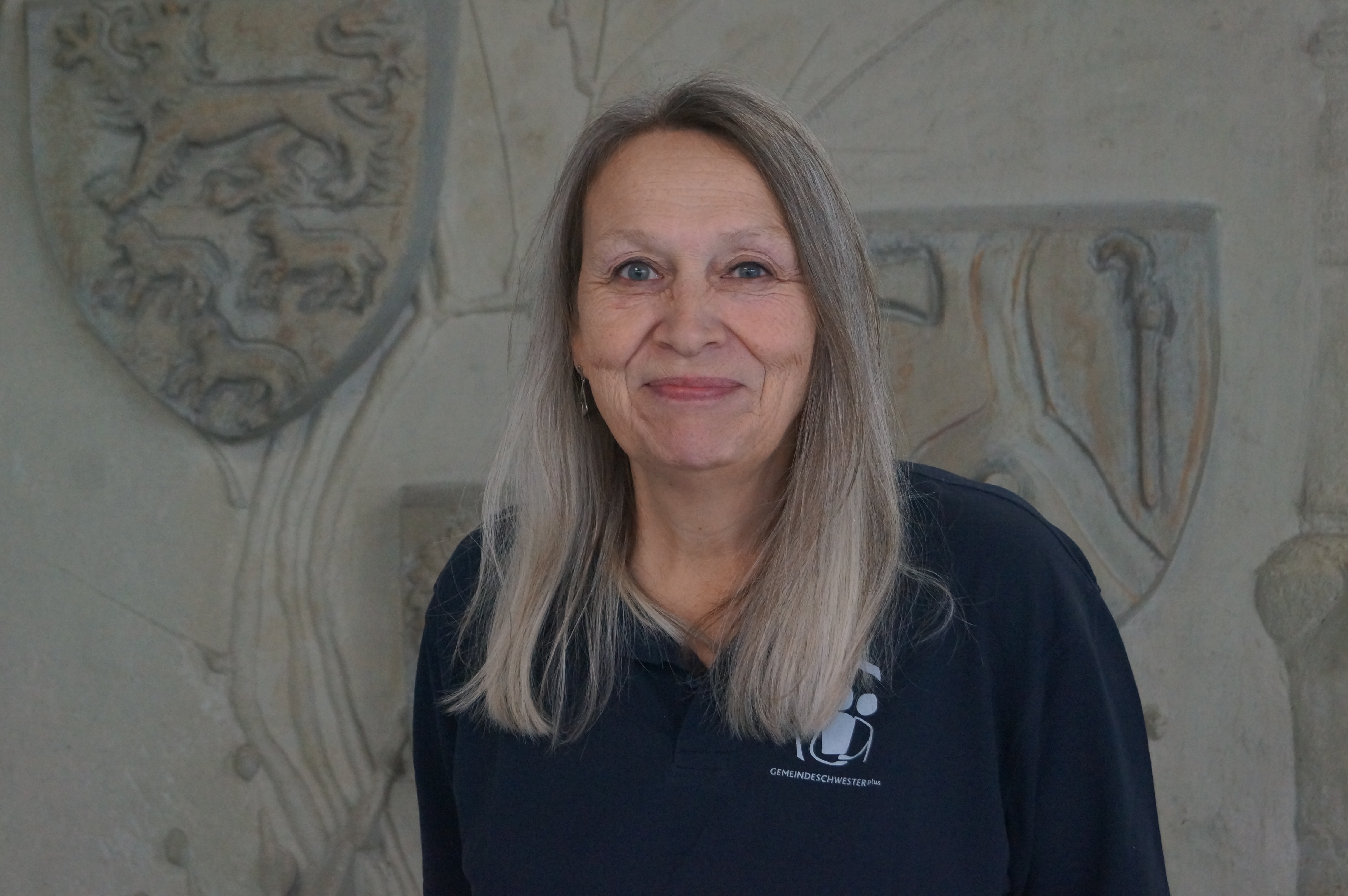Eine Frau mit langen grauen Haaren vor eine steinernen Wand.