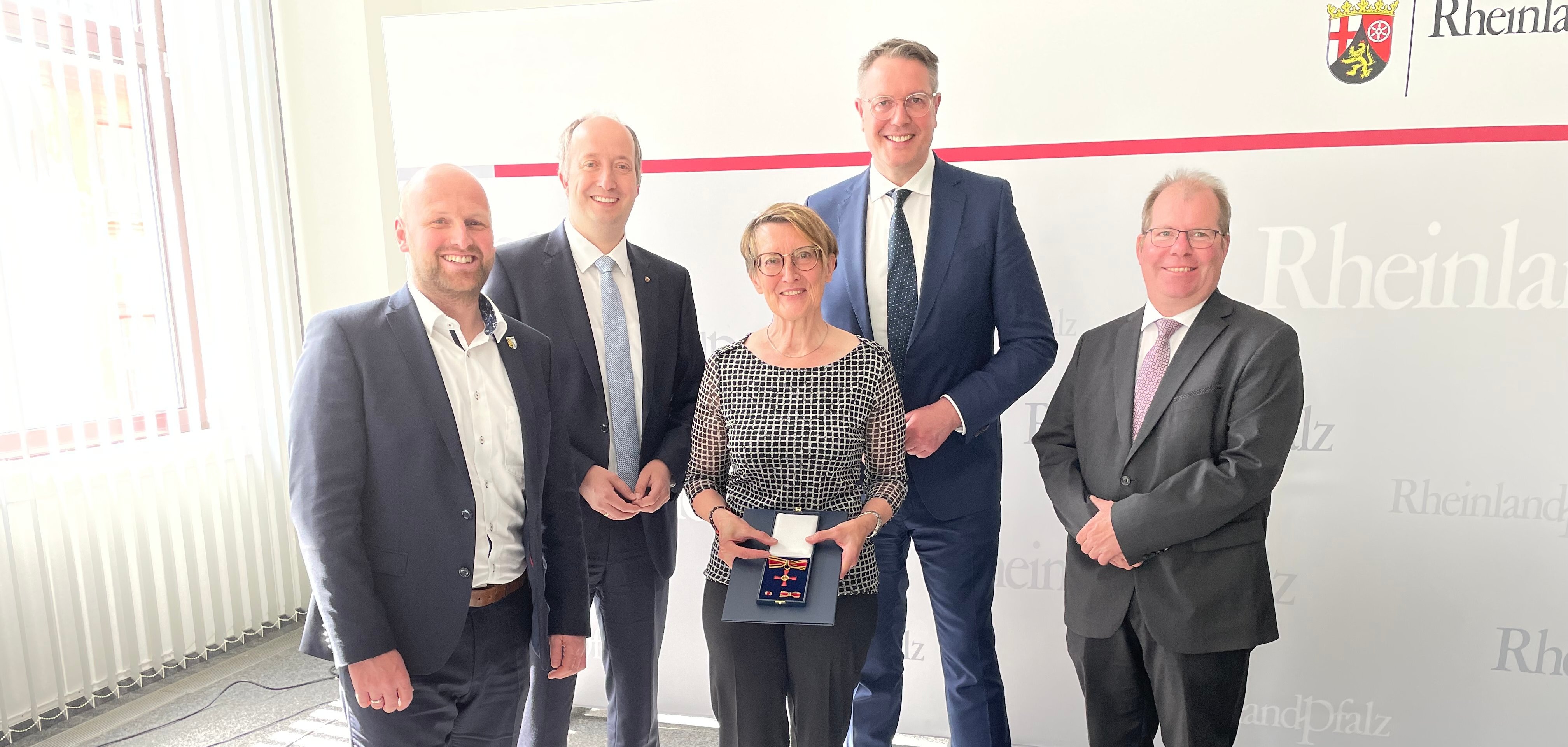 Vier Männer in Anzügen stehen um eine Frau, die das Bundesverdienstkreuz in der Hand hält. 