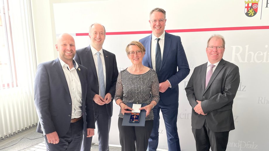 Vier Männer in Anzügen stehen um eine Frau, die das Bundesverdienstkreuz in der Hand hält. 