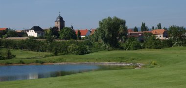 Blick vom Golfgarten auf Dackenheim, im Vordergrund gepflegter Rasen mit See und Fähnchen des Golfplatzes. 