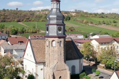Die Kallstadter Kirche von Osten aus aufgenommen. Im Vordergrund der Ort, im Hintergrund Weinberge und Gehölze. 
