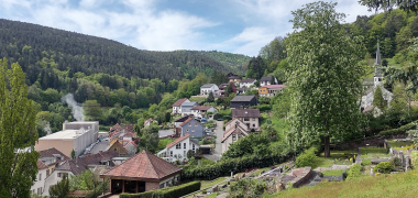 Das im Tal zwischen Hügeln und Wald liegende Dorf vor blauem Himmel mit weißen Wolken. 