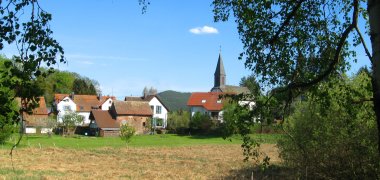 Von Ästen mit Laub umrahmte Dorfansicht mit protestantischer Kirche im Ortsteil Hertlingshausen. 