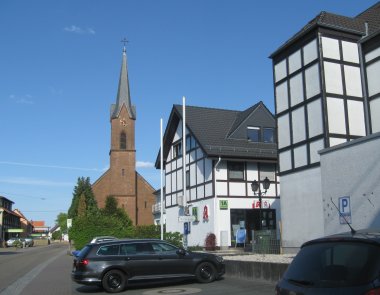 Die katholische Kirche in Carlsberg im hinteren Teil, vorne zwei Fachwerkhäuser und parkende Fahrzeuge. 