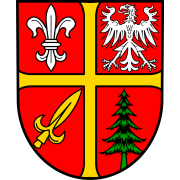 Wappen von Carlsberg. Rot, geviertelt von einem gelben Kreuz. Links oben eine weiße Lilie, rechts oben ein weißer Adler, links unten ein gelbes Schwert, rechts unten ein grüner Tannenbaum. 