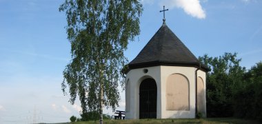 Die Kapelle ist ein kleiner weißer Bau mit rundem Dach auf einem grasbewachsenen Hügel. Daneben ein hoher Baum.