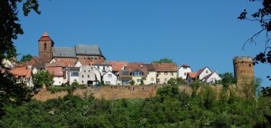 Häuser des Dorfes umgeben von der Befestigungsmauer, rechts ein Turm. Im Vordergrund Bäume. 