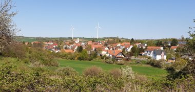 Ortsansicht, im Vordergrund viel grün mit Büschen und Bäumen, dahinter der Ort, im Hintergrund blauer Himmel. 