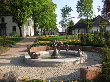 Ein Brunnen in einem gepflasterten Kreis. Dahinter Gebäude und Bäume. 