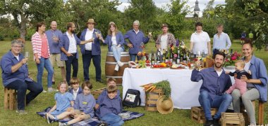 Menschen mit Weingläsern in der Hand, teilweise mit Winzerkitteln bekleidet, stehen und sitzen um einen gedeckten Tisch, . 
